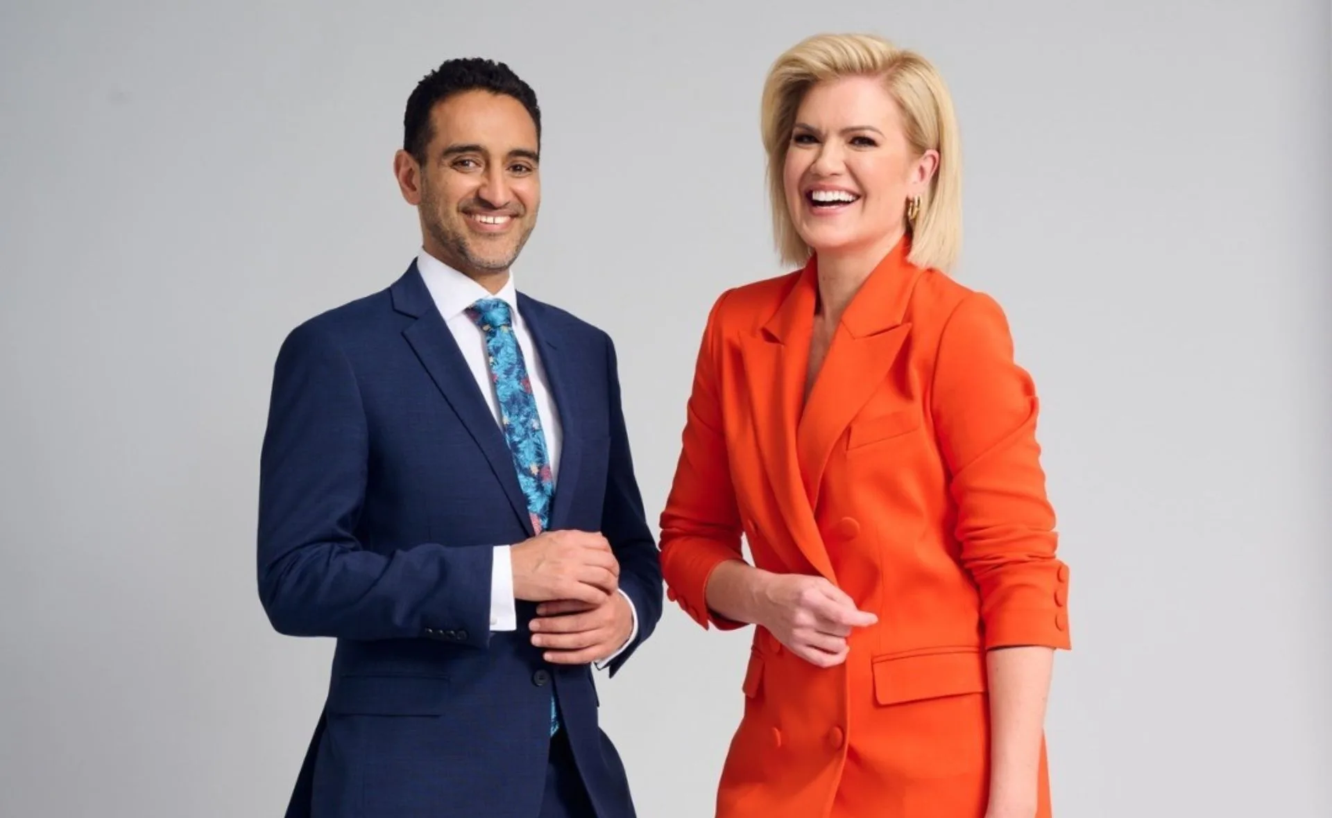 Two smiling hosts stand side by side; the man in a suit and the woman in an orange jacket.