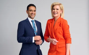 Two smiling hosts stand side by side; the man in a suit and the woman in an orange jacket.
