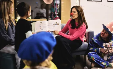 Child reading session with woman in red nose holding a book, surrounded by kids and a person in clown attire holding a toy.