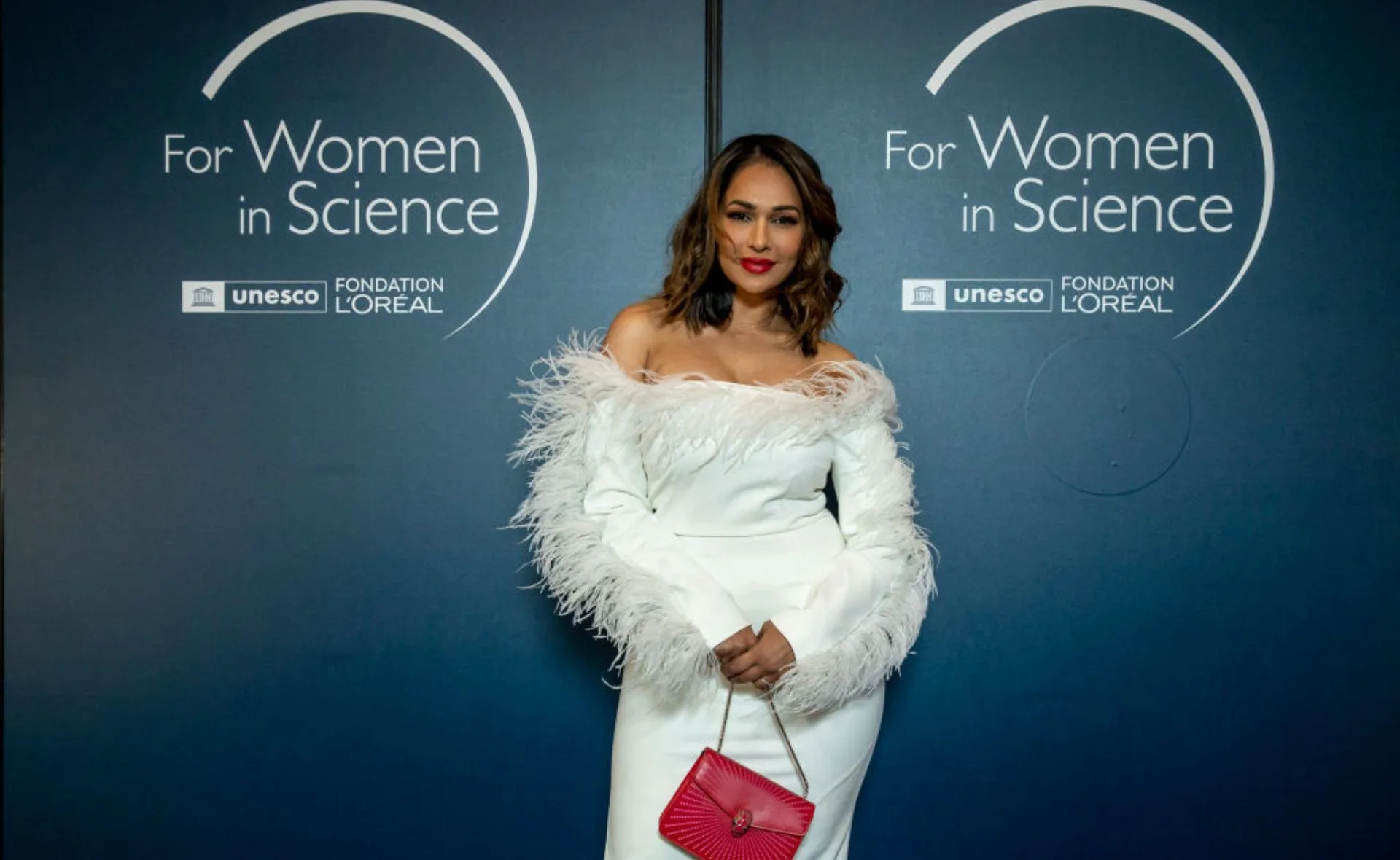 Woman in elegant white dress with red purse at "For Women in Science" event backdrop.