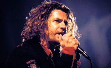 Singer with long curly hair passionately performs on stage, holding a microphone close.