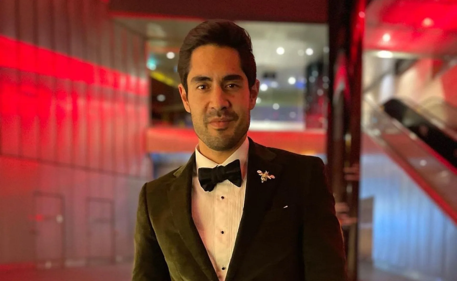 A person in a tuxedo stands in a modern hallway with red lighting, featuring a bow tie and decorative pin.