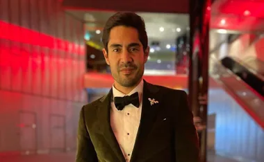 A person in a tuxedo stands in a modern hallway with red lighting, featuring a bow tie and decorative pin.