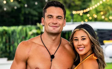 Man and woman smiling together, outdoors, with string lights in background.