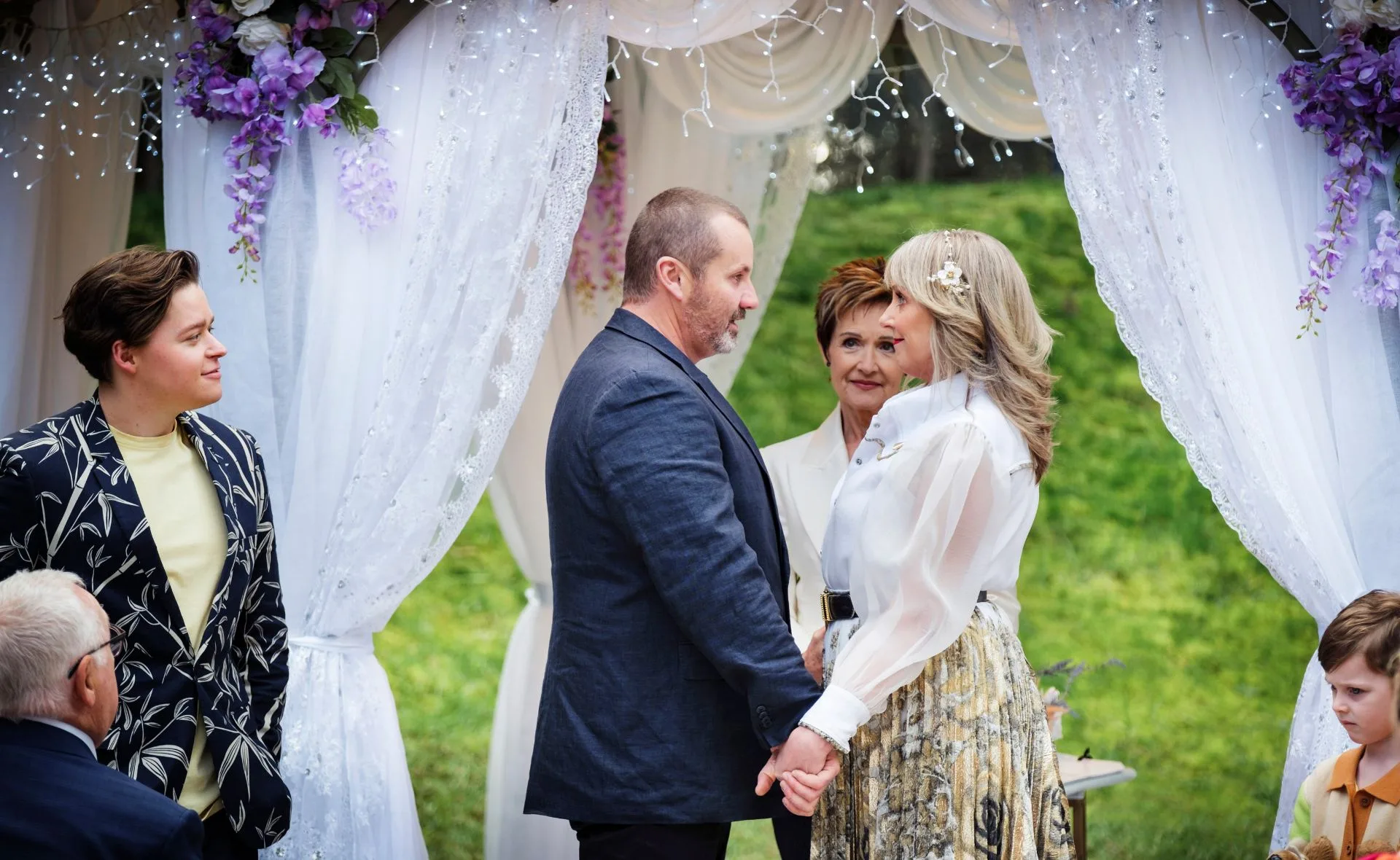 A couple holds hands in a decorated outdoor setting, surrounded by guests and floral drapery.