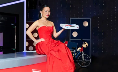 A woman in a red dress holds a pizza box, seated on a counter with a bicycle in the background.