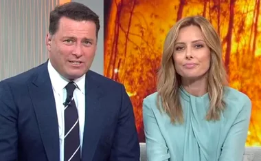 Two news presenters sitting together with a backdrop of orange and red blurred trees.