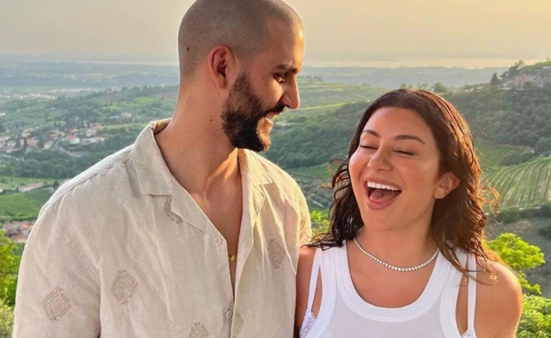 A couple smiling outdoors with a scenic green landscape and sunset in the background.
