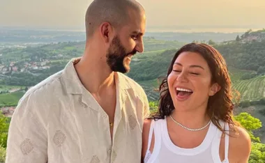 A couple smiling outdoors with a scenic green landscape and sunset in the background.