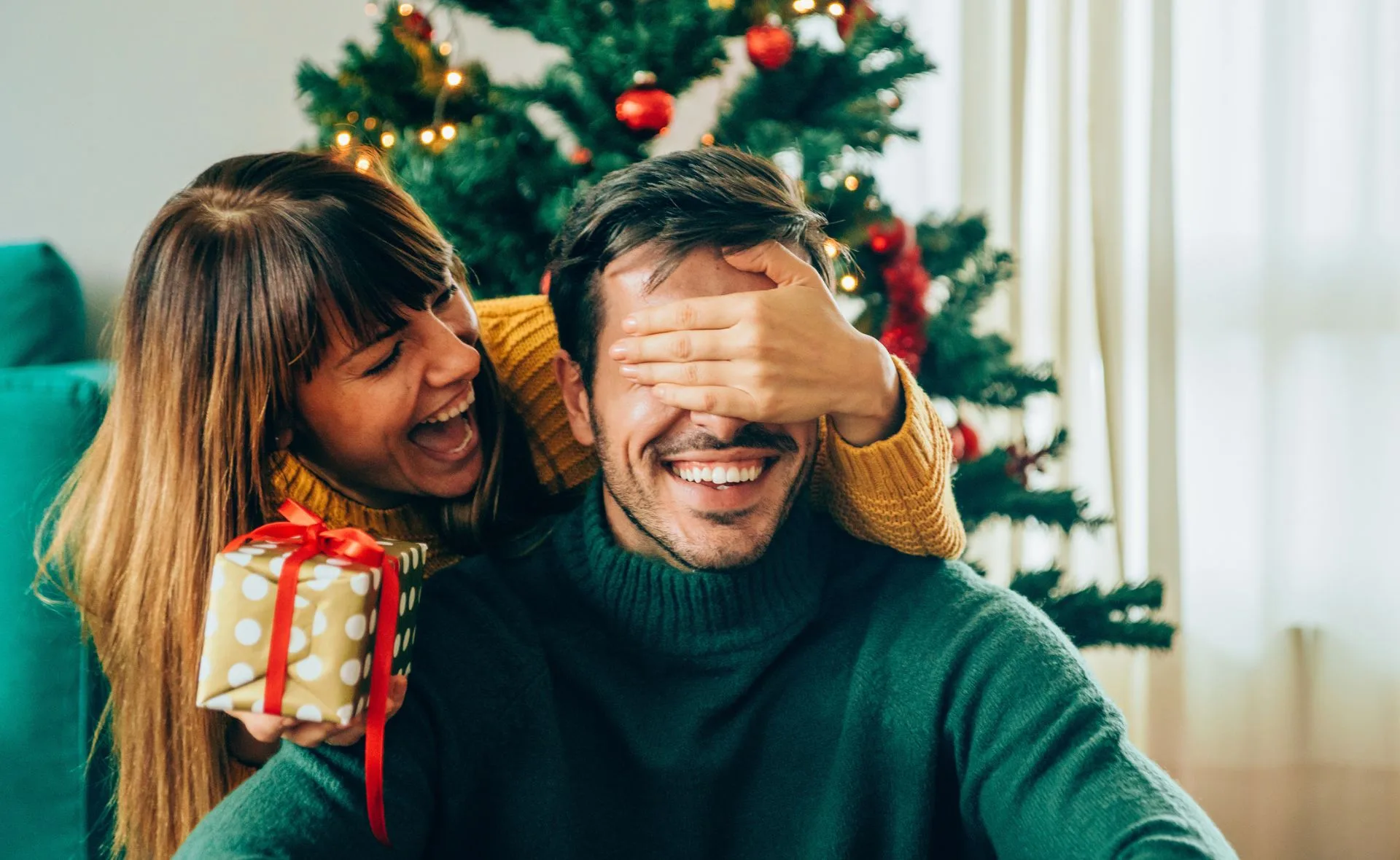 Woman surprises man with gift in front of a Christmas tree, her hand covering his eyes, both smiling joyfully.