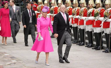 Couple in formal attire, woman in bright pink dress and fascinator, walk past ceremonial guards in red uniforms.