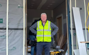 Man in a construction site wearing a high-visibility vest, standing with hands on hips and smiling.