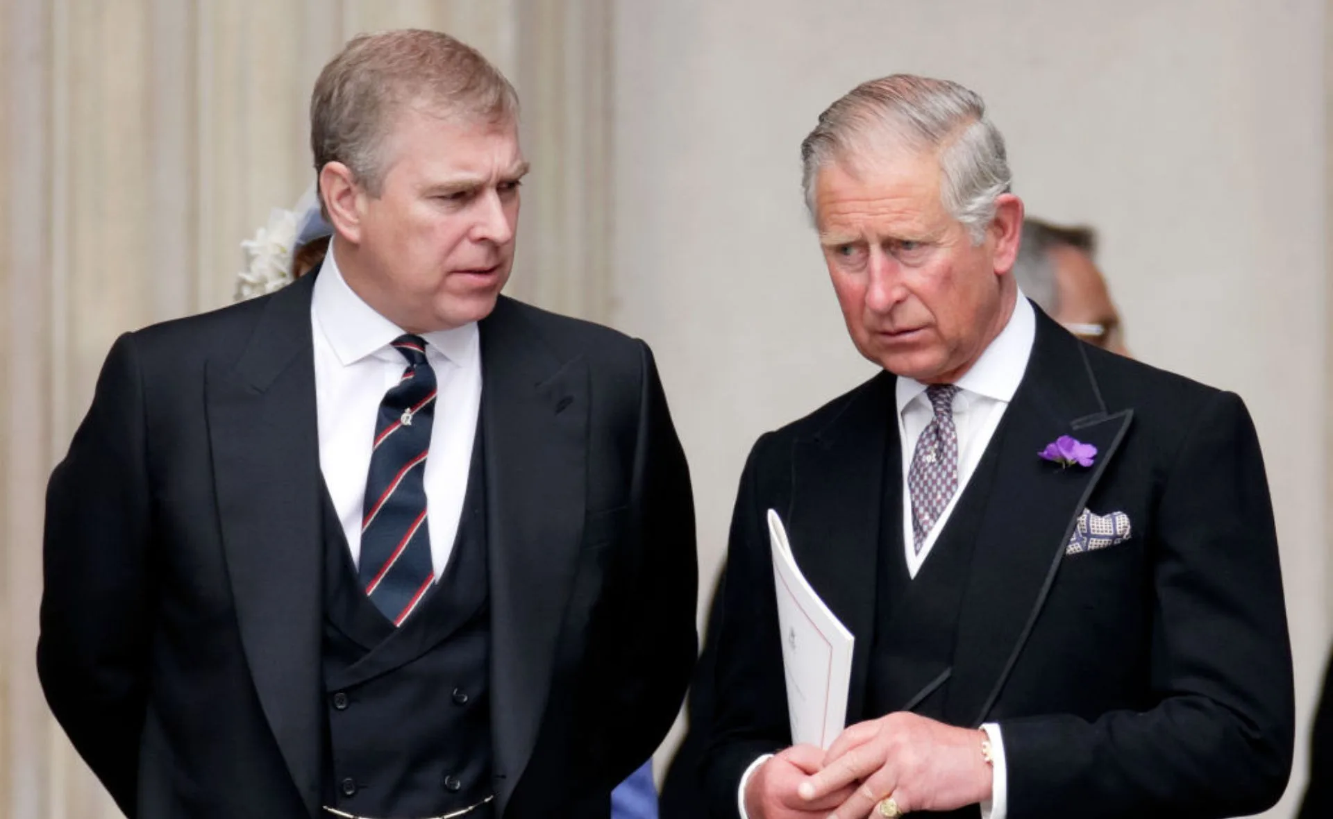 Two men in formal suits standing and conversing, one holding a booklet, outside a building.