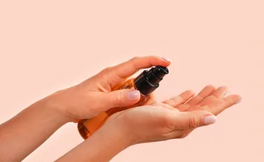Hands applying a clear substance from a pump bottle onto one palm, over a peach-colored background.