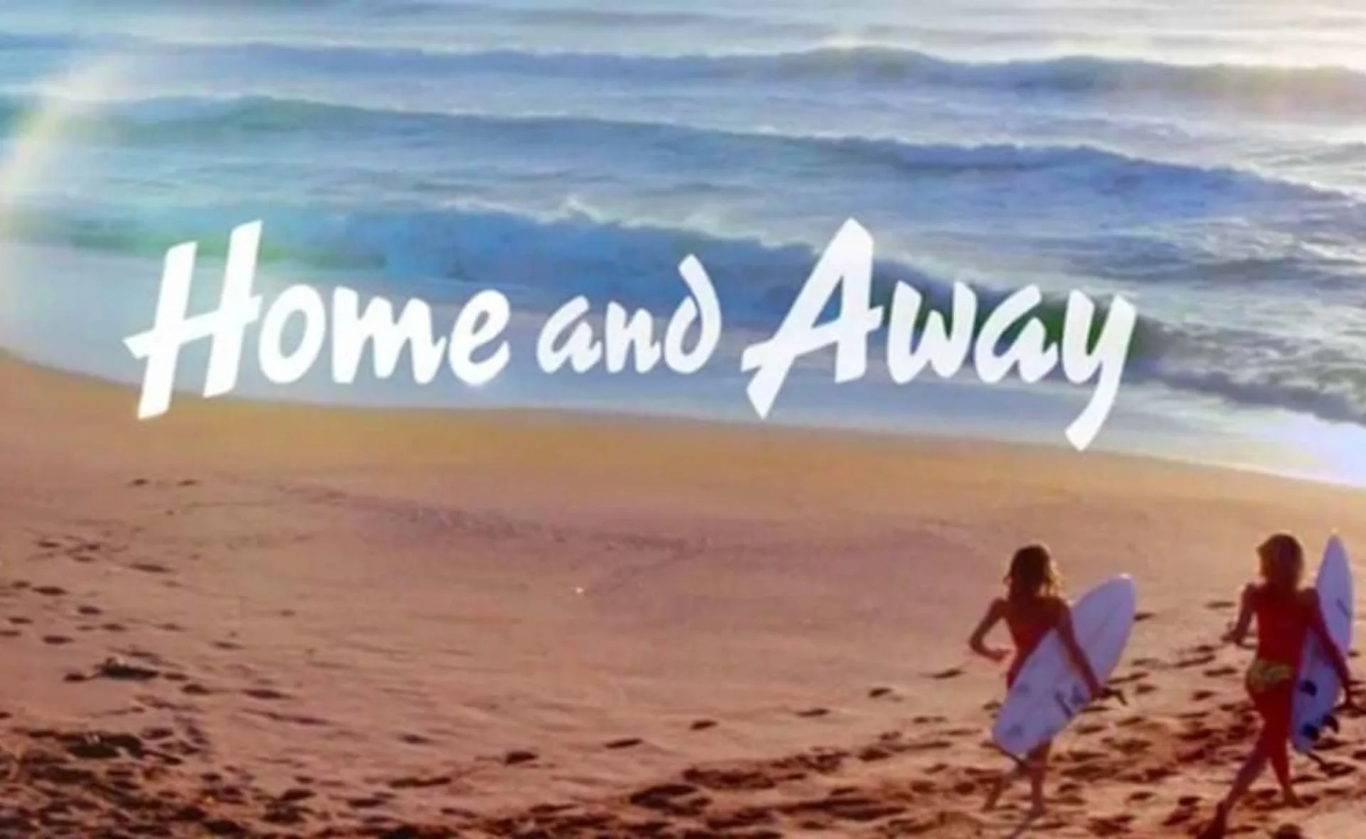 Two surfers walk on a beach with the "Home and Away" logo above them.