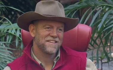 Man in a cowboy hat and red vest smiling, sitting on a red chair, surrounded by greenery.