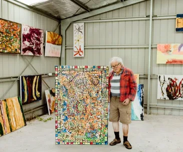 Man in a plaid shirt and shorts stands in an art gallery, showcasing a colorful abstract painting with mosaic elements.