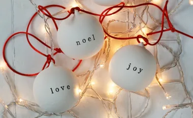 White Christmas baubles with red strings, labeled "love," "noel," and "joy," surrounded by string lights.