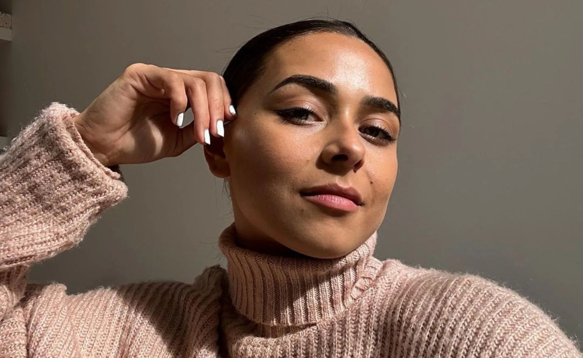 Person in a cozy pink sweater, taking a selfie with a relaxed expression and hand near face.