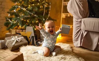 Baby in elephant onesie joyfully holding a gift by a Christmas tree, surrounded by presents and warm lights.