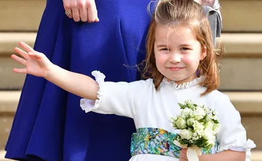 Young girl in a white dress holding flowers, waving, standing beside an adult in a blue dress.