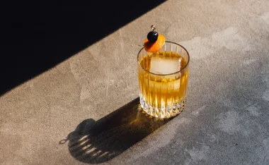A glass of whisky with an ice cube and garnish, casting a shadow on a concrete surface in sunlight.