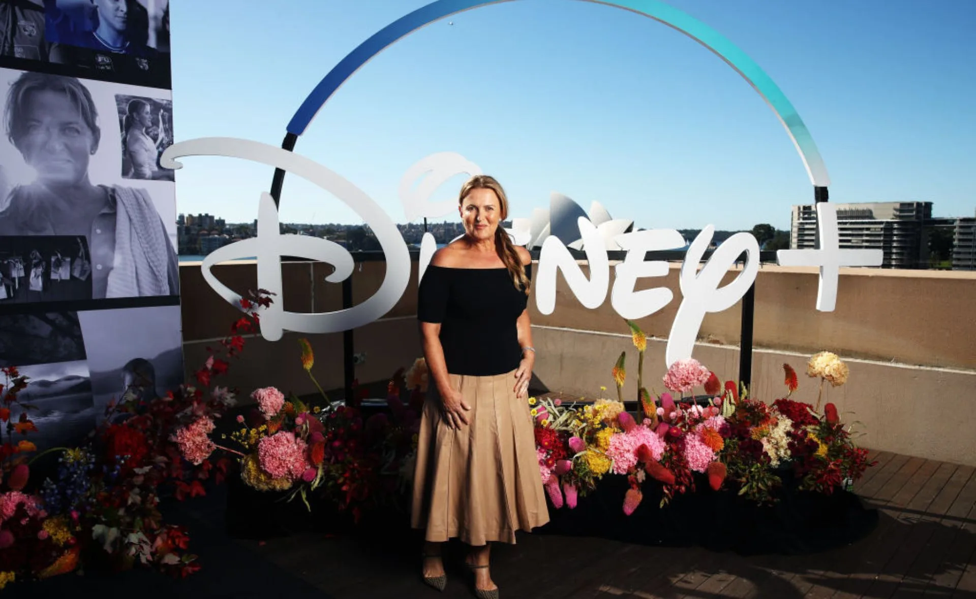 A woman stands in front of a Disney+ sign with colorful flowers, under a clear blue sky.