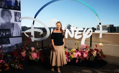 A woman stands in front of a Disney+ sign with colorful flowers, under a clear blue sky.