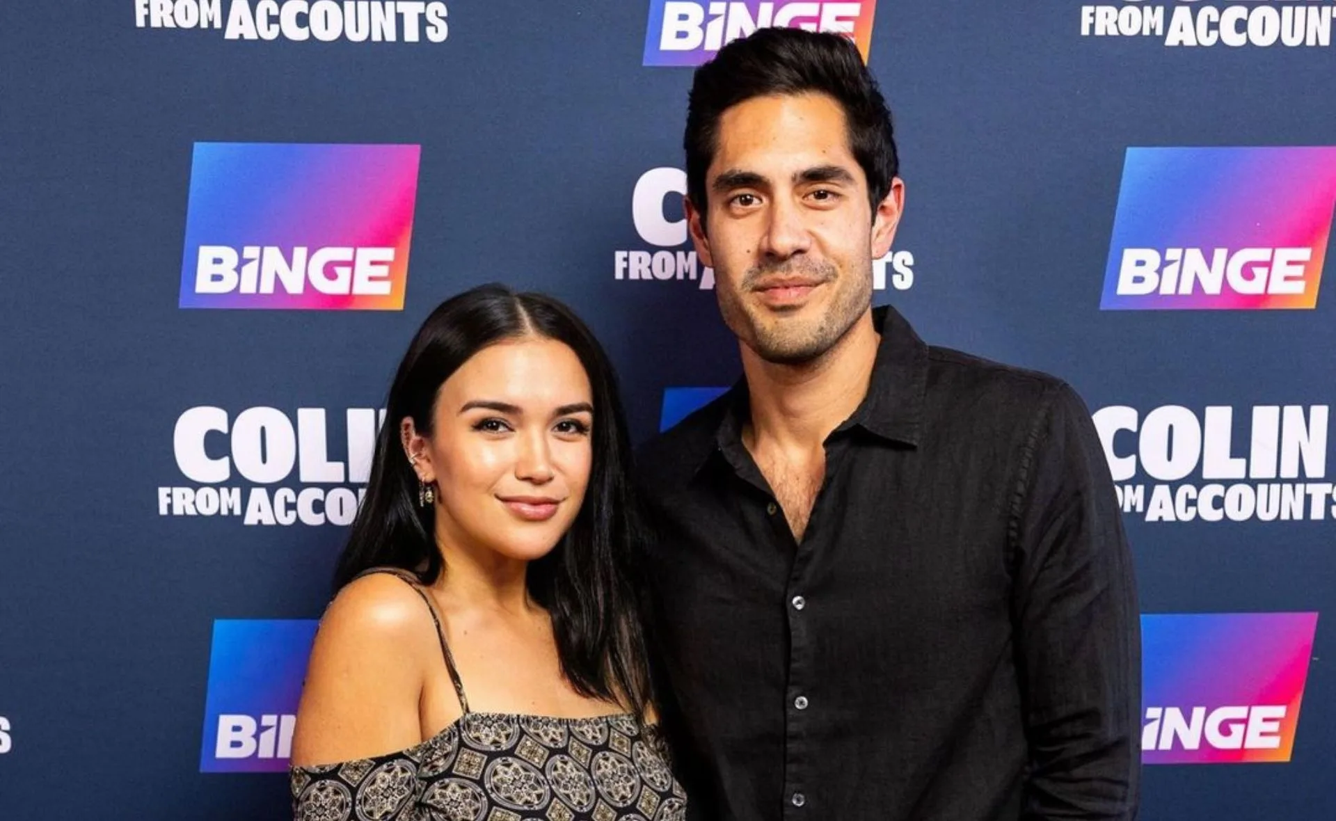 Two individuals pose together at a "Colin From Accounts" promotional event with a Binge backdrop.