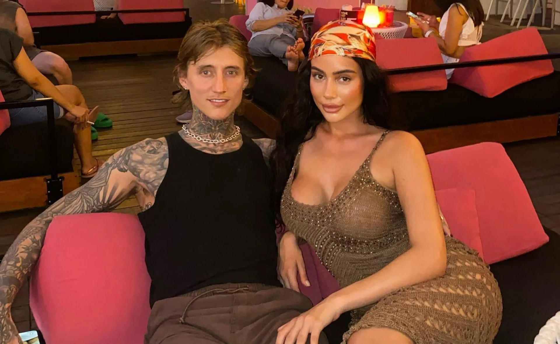 A tattooed man and a woman in a knit dress sit together on pink seats in a lounge setting.
