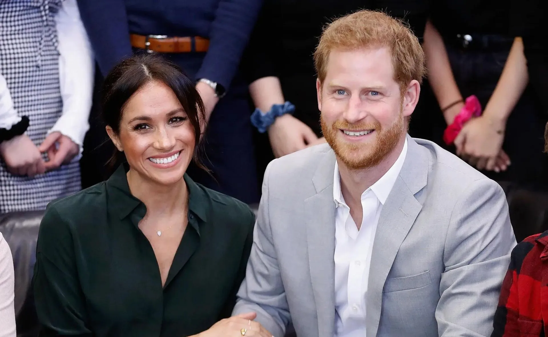 A smiling couple in formal outfits sitting together, surrounded by people in the background.