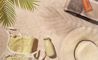 Beach essentials on sandy ground: palm leaf, net bag with towel, sunhat, bottle, folded blanket, sunscreen tube.