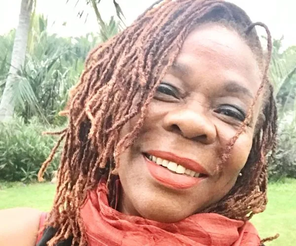 A smiling woman with dreadlocks and a red scarf, outdoors with greenery in the background.