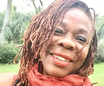 A smiling woman with dreadlocks and a red scarf, outdoors with greenery in the background.