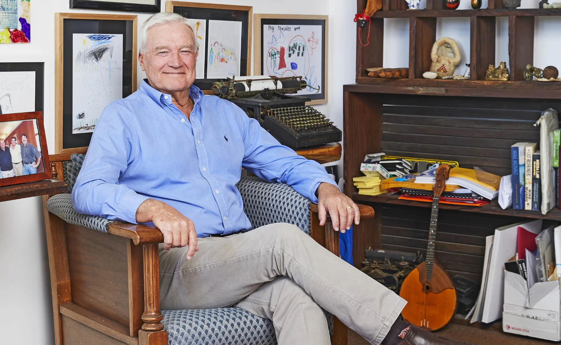 Elderly man in a blue shirt seated in a chair with framed artwork and a vintage typewriter in the background.