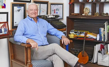 Elderly man in a blue shirt seated in a chair with framed artwork and a vintage typewriter in the background.