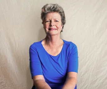 Elderly woman in a blue shirt smiling with a neutral backdrop.