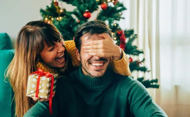 Woman holding gift covers man's eyes, both smiling in front of Christmas tree.