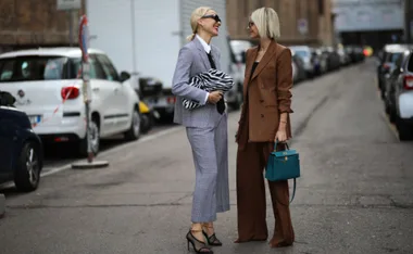 Two women in stylish suits standing on a city street, smiling at each other.