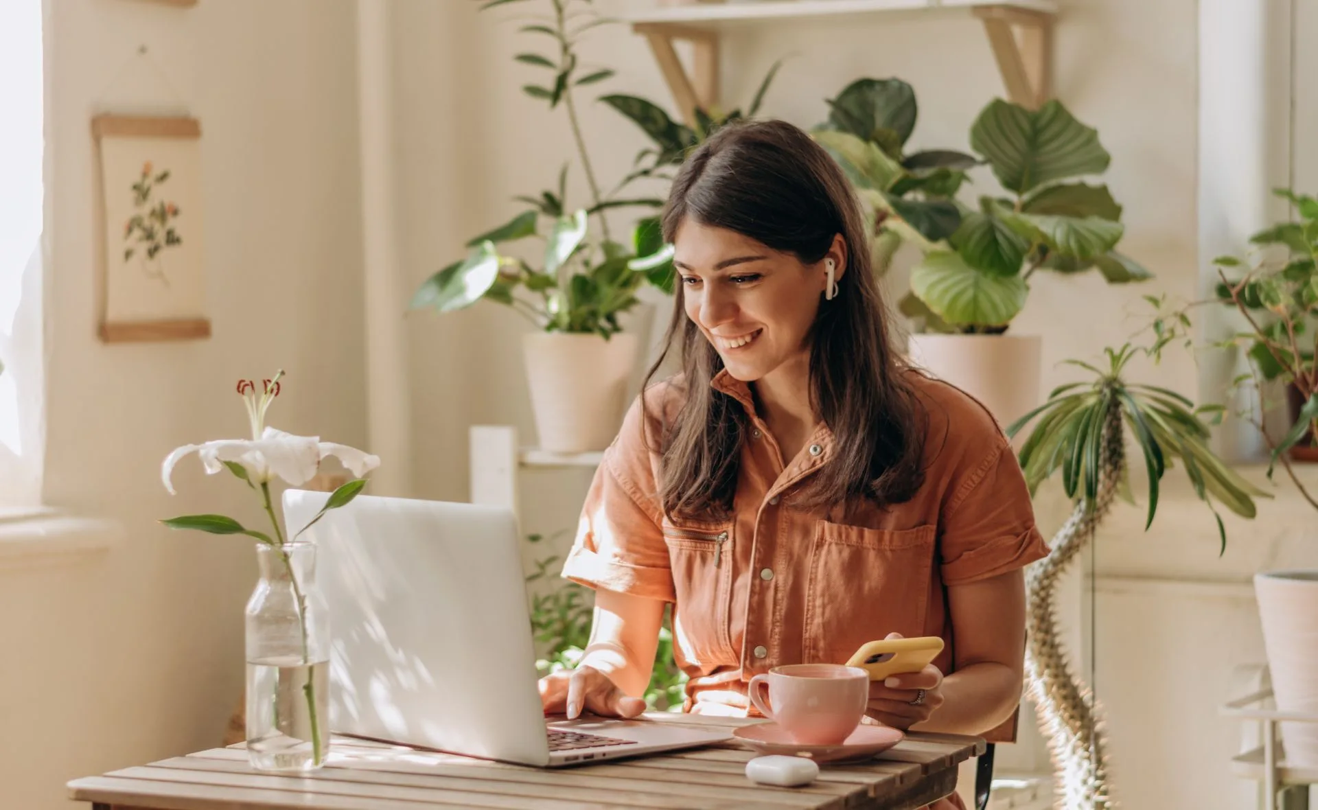 Woman in a cozy home office with plants, using a laptop, holding a phone, and wearing earbuds.