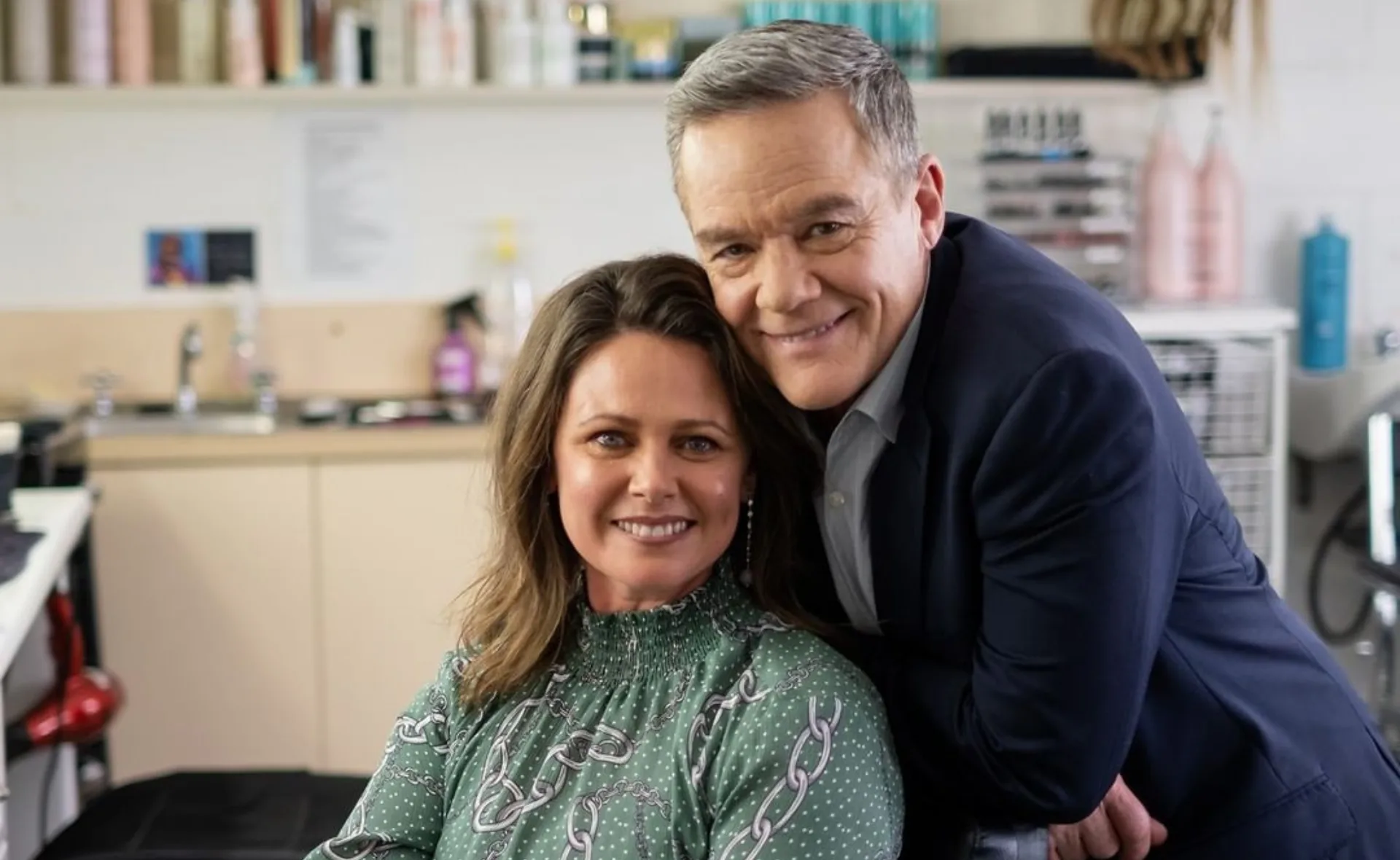 A person with a green patterned blouse seated, smiling, with a man in a suit leaning over, both in a room with shelves.