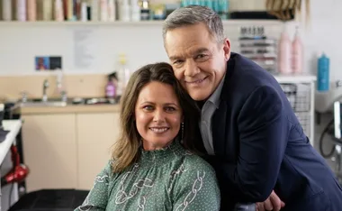 A person with a green patterned blouse seated, smiling, with a man in a suit leaning over, both in a room with shelves.