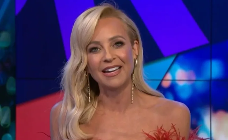 A woman with blonde hair smiles, wearing a strapless red dress and dangling earrings against a colorful studio backdrop.