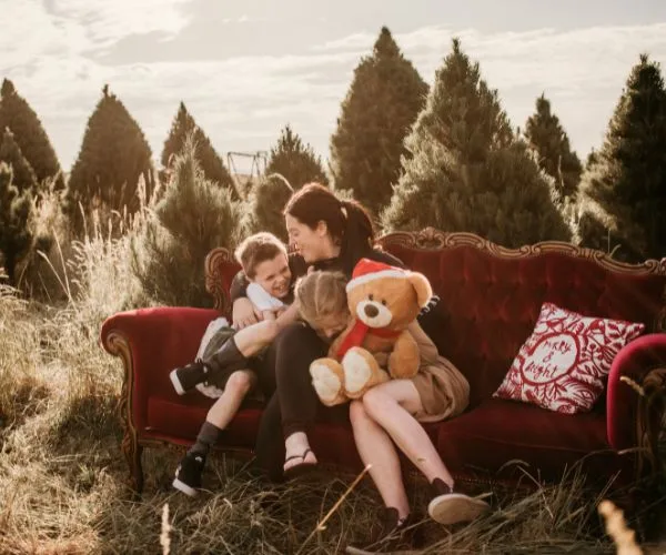 Family on a red sofa outdoors, surrounded by Christmas trees; kids hugging a teddy bear and an adult.