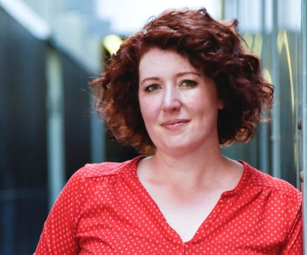 Portrait of a person with curly hair wearing a red shirt, standing in an alley with a soft smile.