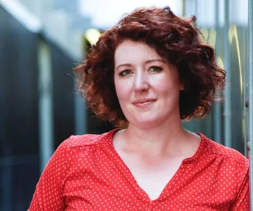 Portrait of a person with curly hair wearing a red shirt, standing in an alley with a soft smile.
