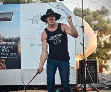 Man wearing a hat and sleeveless shirt performing a whip-cracking act outdoors near a trailer.