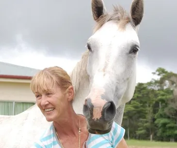 A person smiling beside a white horse, outdoors with a cloudy sky and trees in the background.