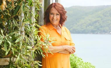 Woman in orange shirt smiling, standing by greenery with a scenic view of the water and hills in the background.