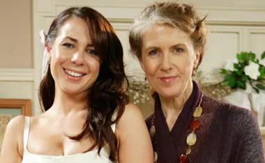 Two women smiling, standing indoors; one is wearing a white dress. The setting is likely from "Home and Away.”